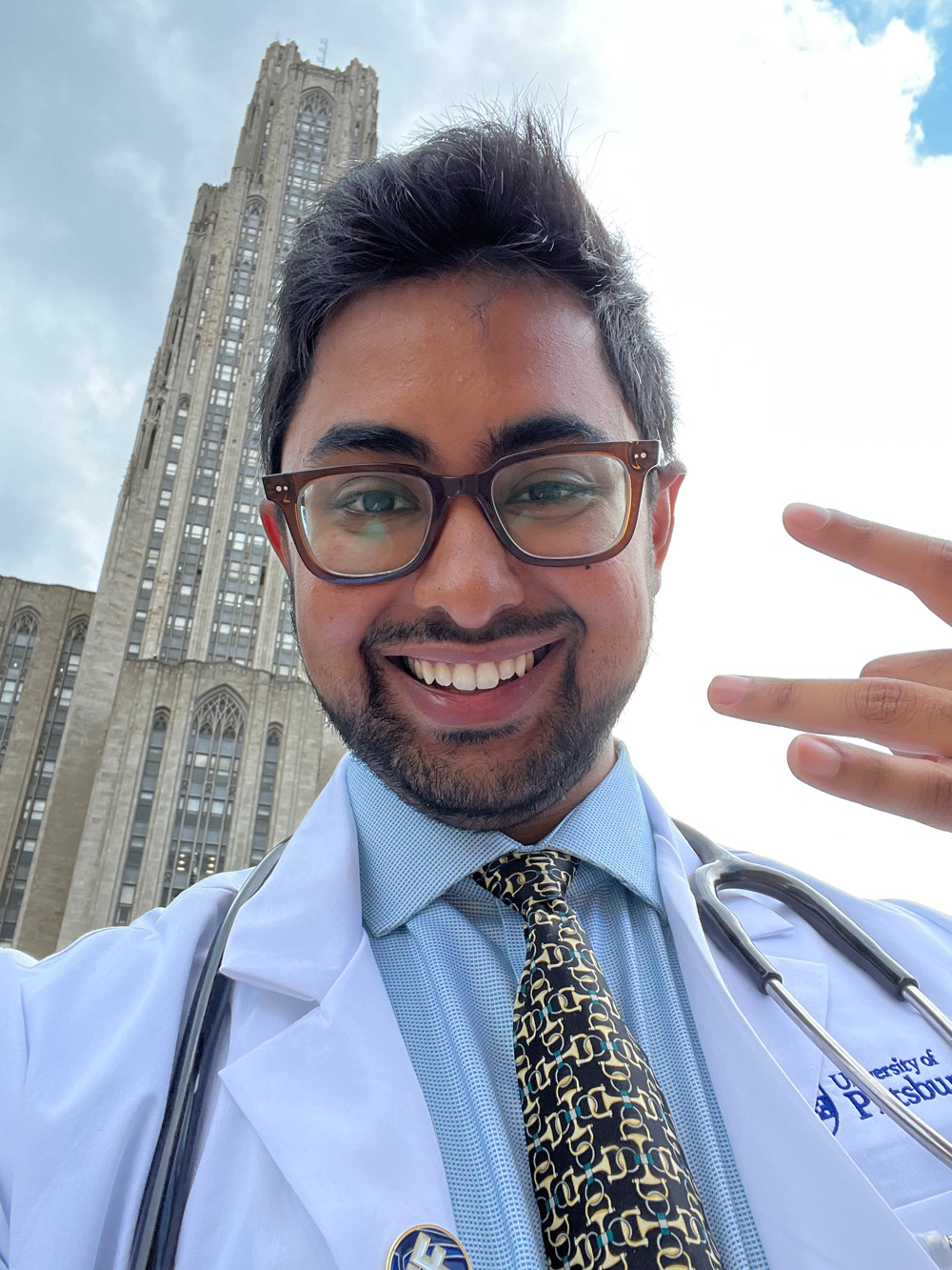 Alum Razeen Khan at the University of Pittsburgh Medical School, wearing his coat and stethoscope, with the Cathedral of Learning in the background.
